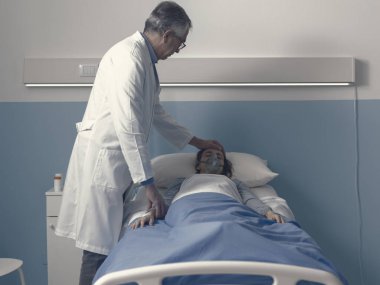 Profesyonel doktor genç bir hastaya hastanede yatarken yardım ediyor. Alnına dokunuyor. Sağlık sigortası kavramına.