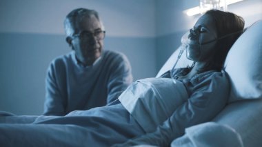 Hastanede kızıyla yan yana oturan ve geceleri ona yardım eden yaşlı adam uyuyor ve oksijen maskesi takıyor.