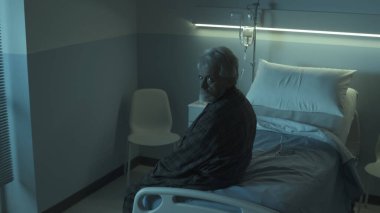 Bunalıma girmiş yaşlı bir adam gece bir hastane yatağında oturmuş kameraya bakıyor.