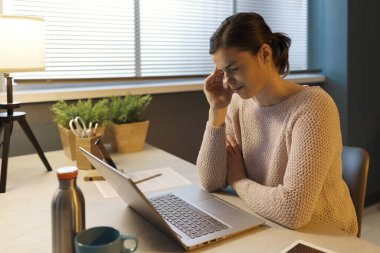 Masasında oturup dizüstü bilgisayarıyla çalışmaktan yorgun düşmüş genç bir kadın kötü bir baş ağrısı çekiyor. 