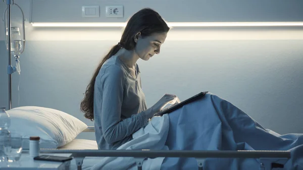 Young smiling sleepless patient lying in a hospital bed at night and connecting with a digital tablet