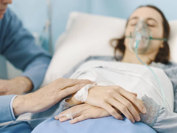 Caring concerned father assisting her sick daughter at the hospital and holding her hand, she is lying in bed and wearing an oxygen mask