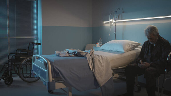 Lonely senior man sitting next to his bed at hospital and making a phone call