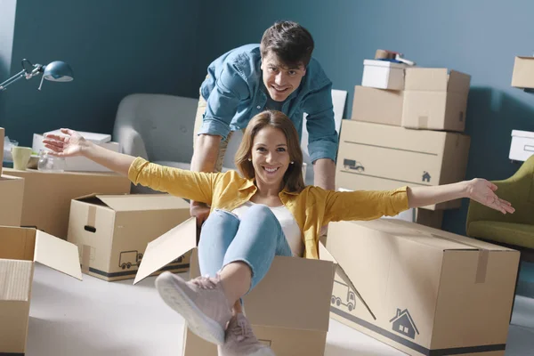 Cheerful happy young couple moving in their new house: she is sitting in a box and her boyfriend is pushing her