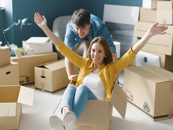 Cheerful happy young couple moving in their new house: she is sitting in a box and her boyfriend is pushing her