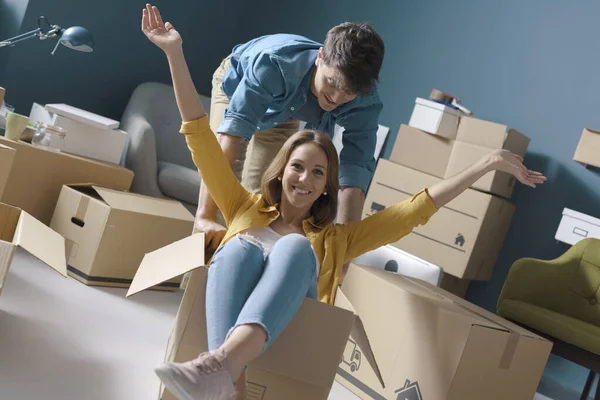 Cheerful happy young couple moving in their new house: she is sitting in a box and her boyfriend is pushing her