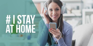 Akıllı telefonuyla müzik dinleyen ve gülümseyen bir kadın: Coronavirus önleme için sosyal medya kampanyasında evde kalıyorum