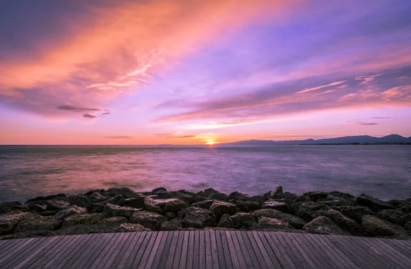 Colorful ultra violet dark sunset over the sea and with a wooden walkway and rocks on the foreground.