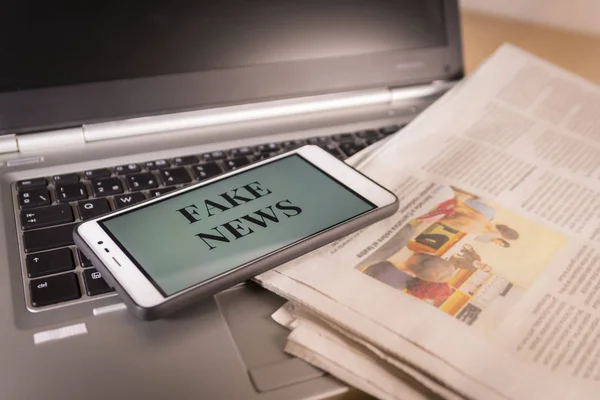 Smartphone mit Fake-News-Wörtern auf dem Bildschirm über einer Zeitung und einem Laptop. Fake News, Falschmeldung — Stockfoto