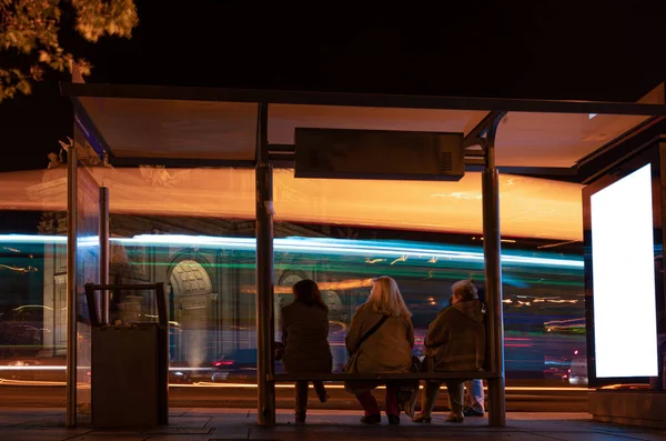 Група людей, які чекають громадського транспорту на нічному автобусному вокзалі з білбордом. Білий порожній екран — стокове фото