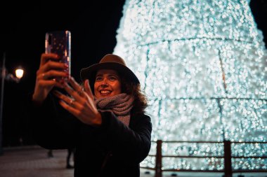 Güzel kıvırcık esmer kadın Madrid 'in arka planında aydınlatılmış bir Noel ağacıyla selfie çekiyor..