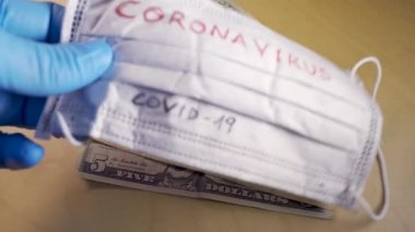 Hands with blue medical gloves putting a respirator mask with the word coronavirus on top of dollar bills