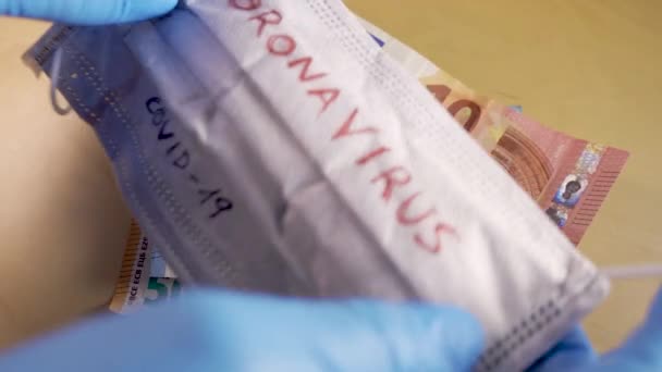 Manos con guantes médicos azules poniendo una máscara respiratoria con la palabra coronavirus encima de los billetes de euro — Vídeos de Stock