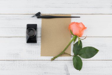 Beyaz ahşap zemin üzerinde turuncu gül boş kahverengi kart, eğik kalem ve mürekkep şişe süslenmiş 