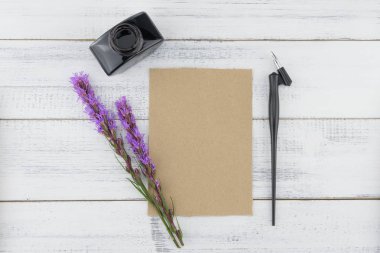Boş kahverengi kart, eğik kalem ve mürekkep şişe beyaz ahşap zemin üzerinde mor liatris çiçeklerle dekore edilmiş