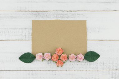 Boş kahverengi kart pembe Gül kağıt çiçeklerle süslemek ve Beyaz ahşap zemin üzerine yeşil yaprakları