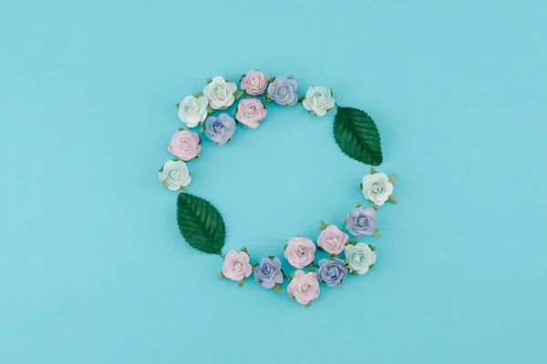 Round wreath made from blue tone paper flowers and green leaves on pastel blue background with copy space