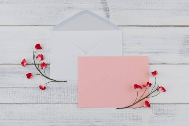 Beyaz ahşap zemin üzerinde kırmızı kağıt çiçeklerle dekore boş pembe kart ve beyaz zarf 