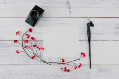 Beyaz ahşap zemin üzerinde kırmızı kağıt çiçeklerle boş beyaz kartı, eğik kalem ve mürekkep şişe süslemeleri