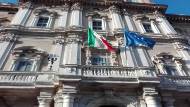 İtalyan ve Avrupa Birliği bayrakları, tarihi hükümet binası cephesinin önünde rüzgarda birlikte sallanıyor.