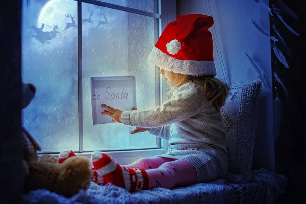 Cute little girl sitting by the window with a letter to Santa Claus