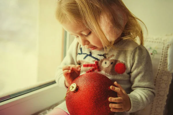 Niña en Navidad con decoraciones de Navidad bola roja sentado en una ventana — Foto de Stock