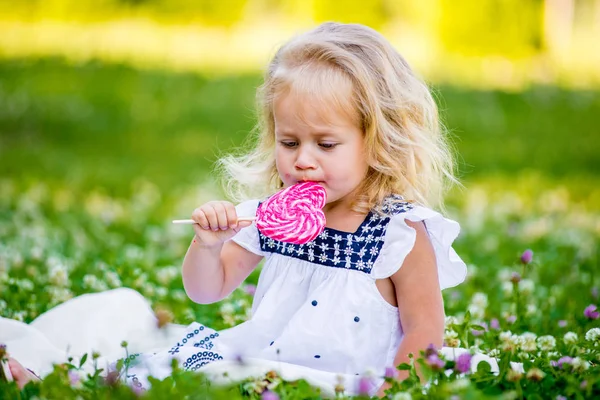 Gelukkig klein meisje eten van een stukje van snoep op een stokje in de vorm van hart. — Stockfoto