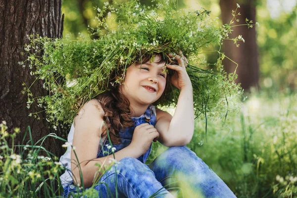 Gelukkig meisje met een krans op een hoofd zitten door een boom in het bos. — Stockfoto