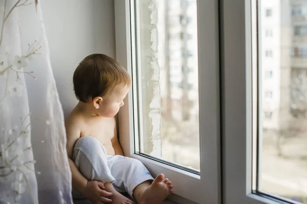 Un niño sentado junto a la ventana con un osito de peluche en las manos — Foto de Stock