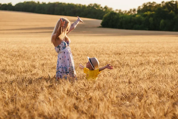 Mooie jonge moeder wandelen met haar jonge zoon op het tarweveld. Moeder en zoon hebben hun handen in de lucht bij zonsondergang — Stockfoto