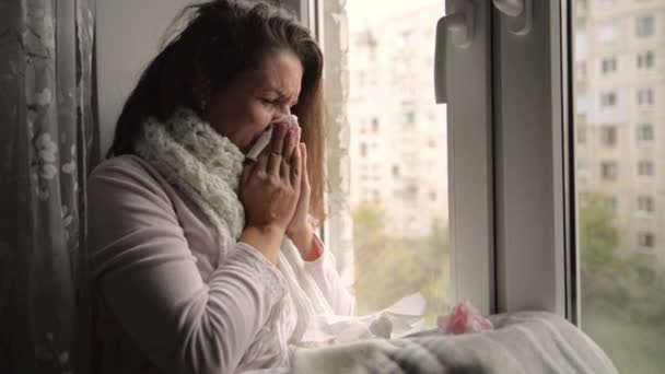 En kall, kylda kvinna nyser. Flicka som blåser näsan sitter vid fönstret. — Stockvideo