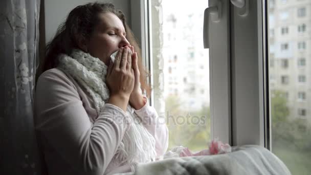 A cold, chilled woman sneezes. Girl blowing his nose while sitting at the window. — Stock Video