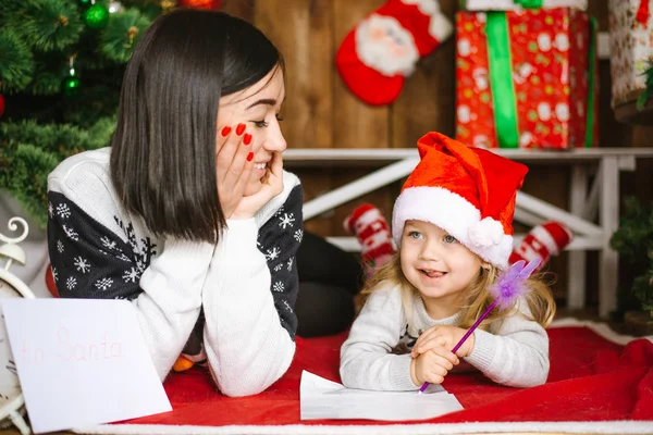 Focus sélectif sur la maman souriante portant une casquette rouge couchée sur le sol près du sapin de Noël avec sa petite fille mignonne écrivant pour le Père Noël — Photo