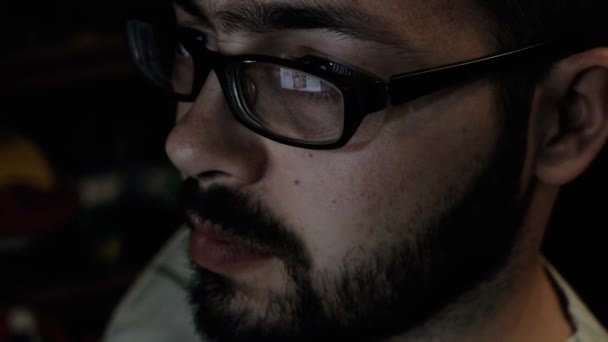 Πορτρέτο ενός γενειοφόρου νεαρού άνδρα που φοράει γυαλιά, κάθεται στο γραφείο του εργάζεται σε έναν υπολογιστή. Οθόνη υπολογιστή αντανακλά τα γυαλιά του. — Αρχείο Βίντεο