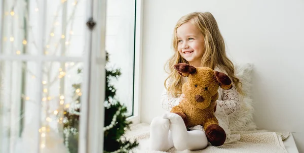 Petite fille avec un cerf en peluche assis sur la fenêtre. Un enfant regarde par la fenêtre et attend Noël, Père Noël — Photo