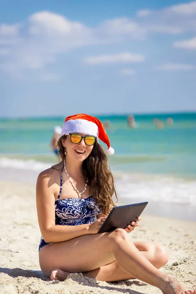 Dziewczyna w Santa Claus kapelusz na plaży z laptopem, tablet — Zdjęcie stockowe