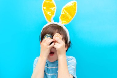 Tavşan kulaklı küçük çocuk mavi arka planda iki boyalı yumurta tutuyor. Paskalya kartı