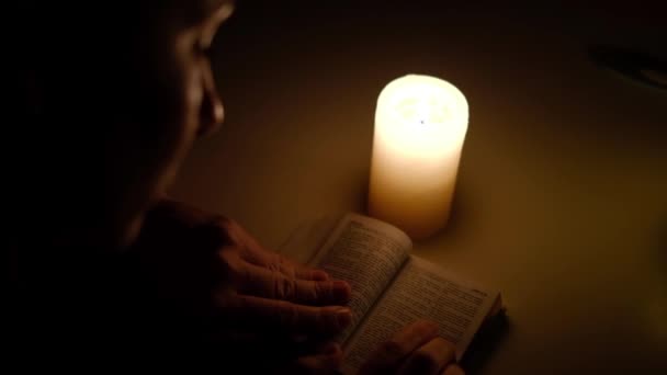 En kvinna sitter och läser en bibel vers i levande ljus på natten, tittar på texten med fingret, närbild — Stockvideo