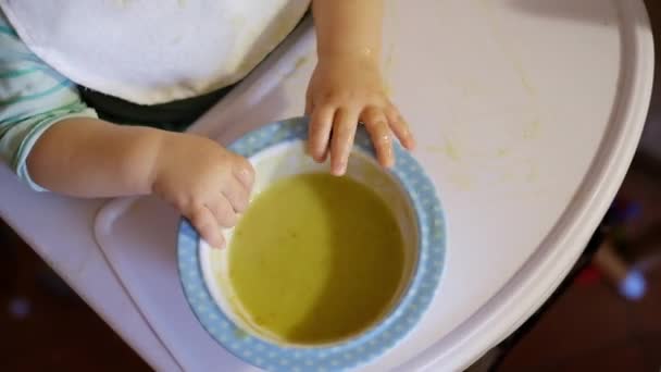 Un niño pequeño come comida para bebés con las manos de un platillo que se derrama en gachas — Vídeos de Stock