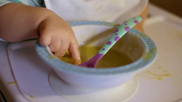 Один маленький ребенок ест детское питание руками от блюдца, разливающегося в кашу. — стоковое видео