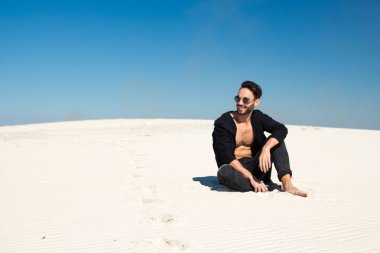 Güneş gözlüğü ve siyah elbiseli bir adam kumsalda oturuyor