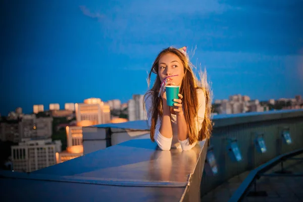 Девушка с бумажной чашкой и трубкой лежит на фоне ночного города — стоковое фото