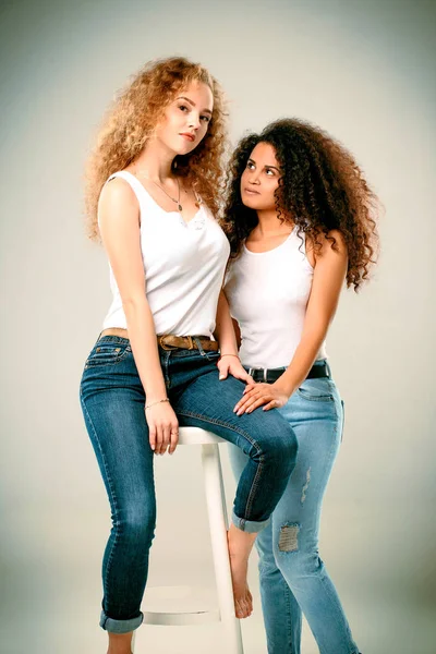 Loira e afro-americana estão sentados em uma cadeira de jeans e camisetas brancas — Fotografia de Stock