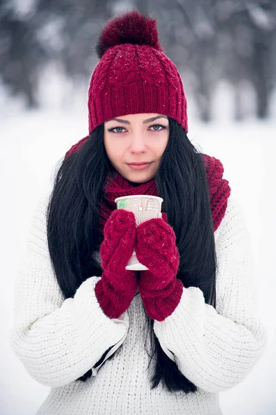 一个漂亮的年轻女子穿着白色毛衣, 戴着一顶明亮的帽子, 围巾和手套, 还有一杯咖啡。 — 图库照片