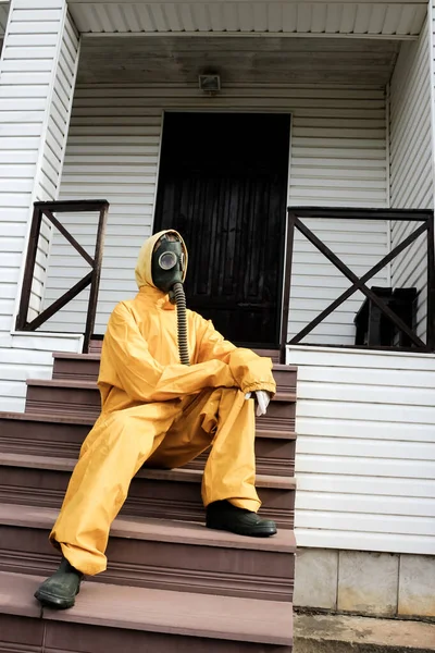 A woman in a gas mask with flexible rubber breathing tube and in yellow protective coverall sitting on the stairs of the house. Environment pollution and self-isolation, quarantine concept