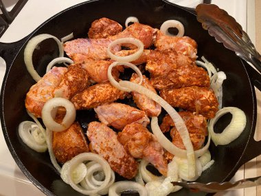 Siyah bir tavada tavuk kanatları ve soğanlar pişiyor.