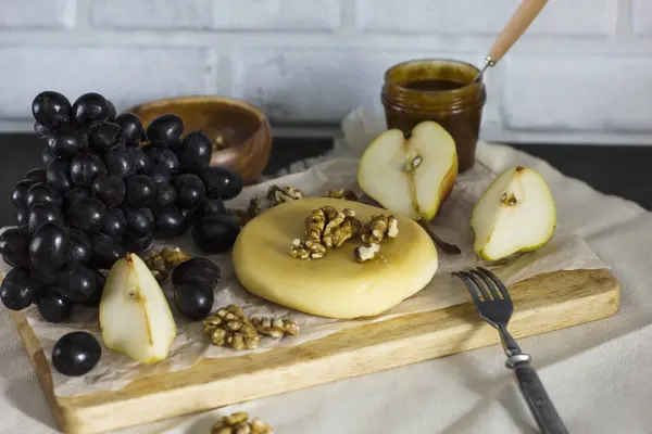 Queijo, mel, uvas, peras e nozes em tábua de madeira na cozinha — Fotografia de Stock