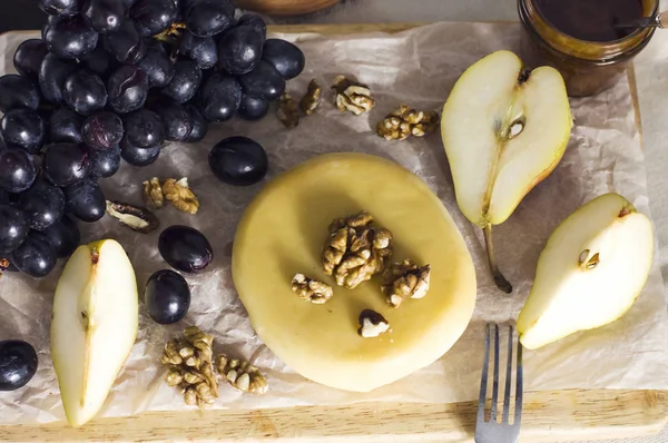 Queijo, mel, uvas, peras e nozes em tábua de madeira na cozinha — Fotografia de Stock