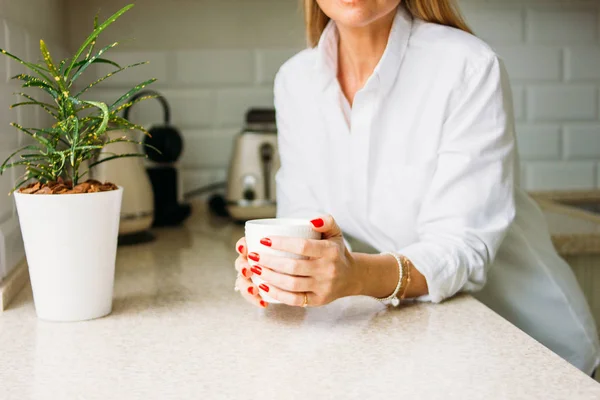 Foto de corte de mulher loira encantadora com cabelo longo e claro em camisa branca com xícara de chá da manhã em mãos na cozinha brilhante antes do dia útil — Fotografia de Stock