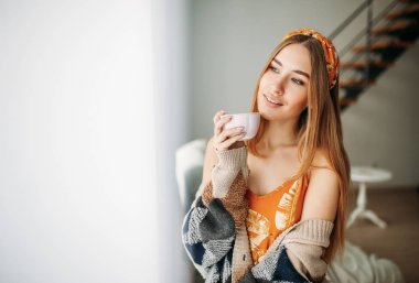 Güzel, gülümseyen, sarışın, uzun saçlı, rahat örülü hırka giyen, evinin penceresinin yanında çay içen bir kız.